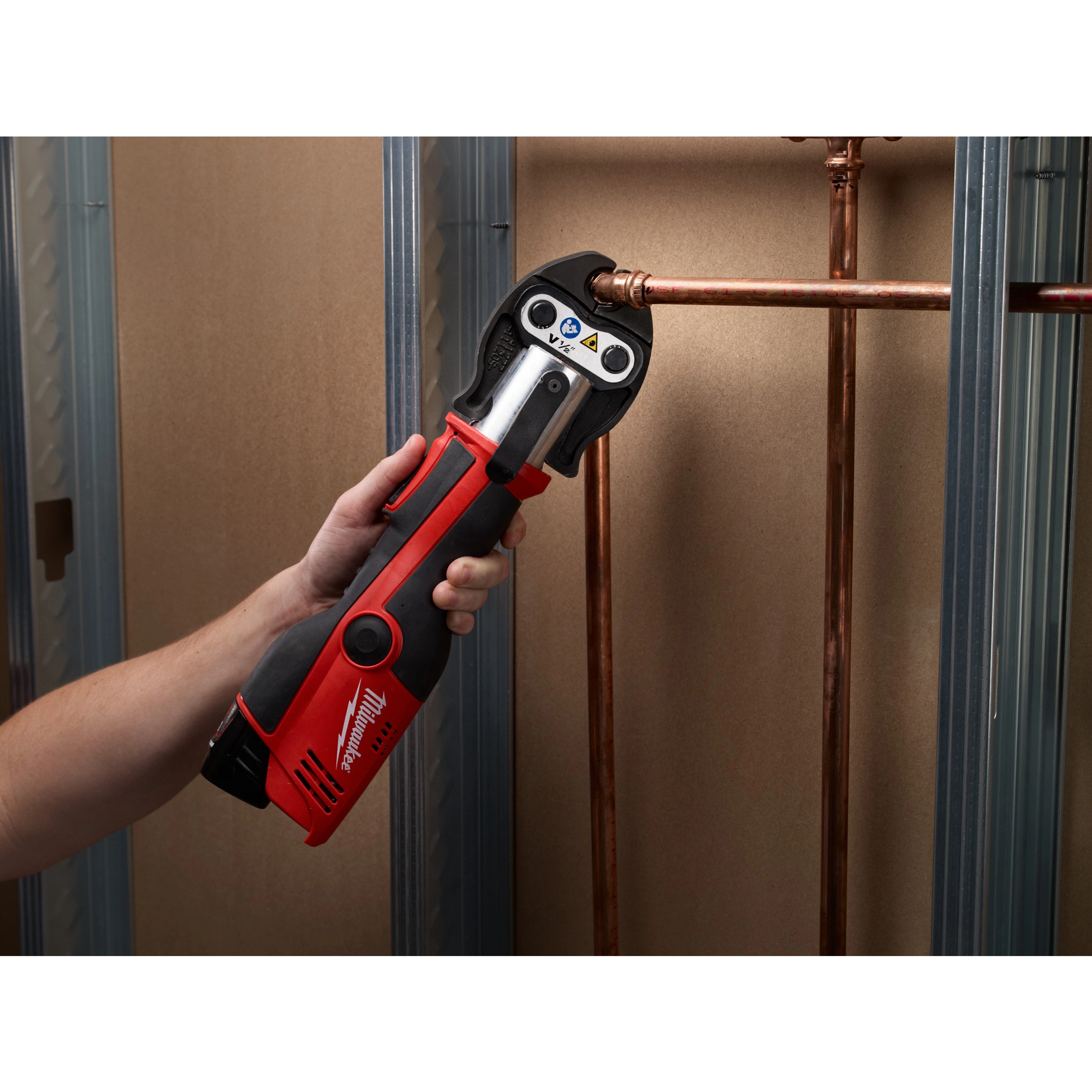 The M12 Force Logic Press Tool Kit, 2473-22, is shown being used to press a copper pipe fitting. The tool is red and black, held by a person's hand, and is pressing a connection in a wall under construction with metal studs.