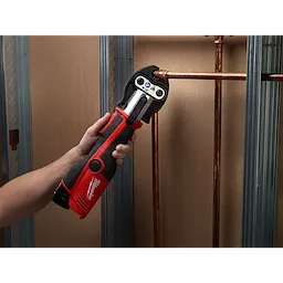 The M12 Force Logic Press Tool Kit, 2473-22, is shown being used to press a copper pipe fitting. The tool is red and black, held by a person's hand, and is pressing a connection in a wall under construction with metal studs.