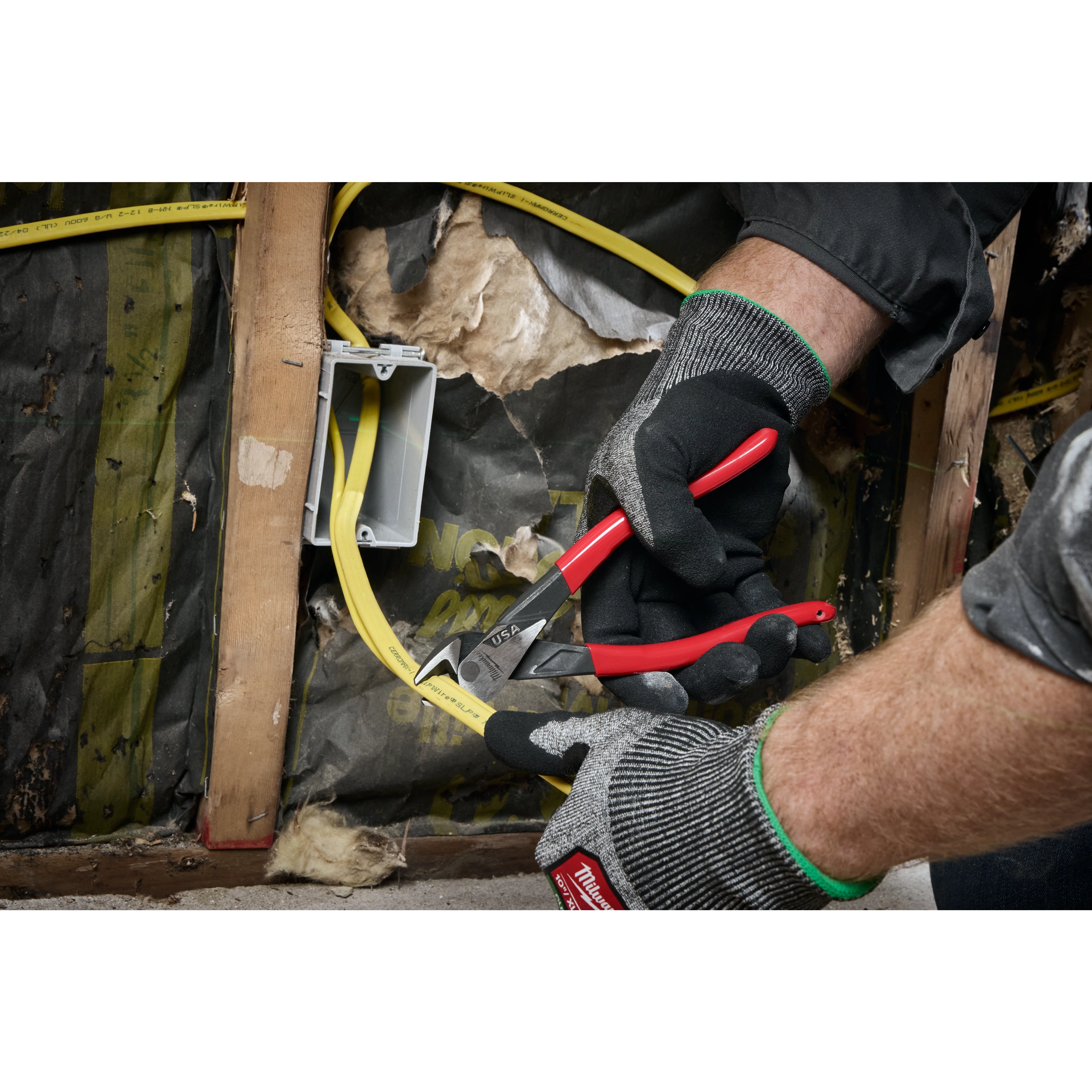 A pair of hands wearing Milwaukee work gloves uses Milwaukee cutting pliers with red and black handles to cut a yellow electrical wire near an electrical box inside a building wall.