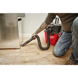 A person uses a vacuum cleaner with a 1-7/8” x 32” to 102” Expandable Hose to clean debris from a hardwood floor next to a cabinet.