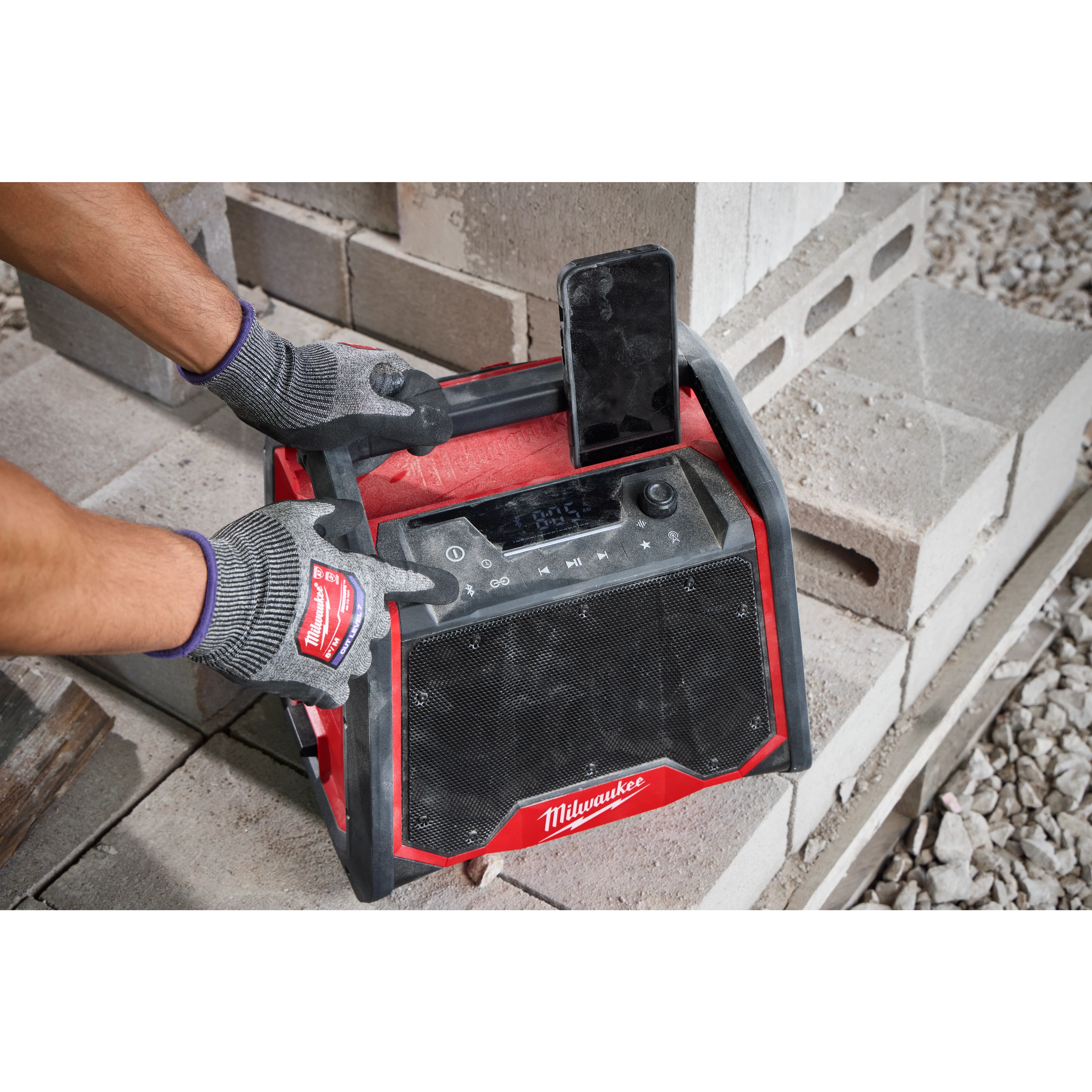 Worker wearing gloves adjusts the M18™ BLUETOOTH® Jobsite Radio + Charger, which is designed with a rugged exterior and large speaker. An upright smartphone is docked on top of the radio. The radio is placed on stacked concrete blocks.