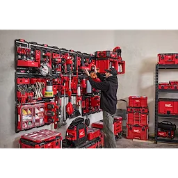 A man organizes tools using the PACKOUT™ Large Wall Plate with multiple shelves and bins. The wall plate and storage units, all in red and black, hold a variety of tools and accessories. The room has additional storage units and a shelf with more toolboxes.