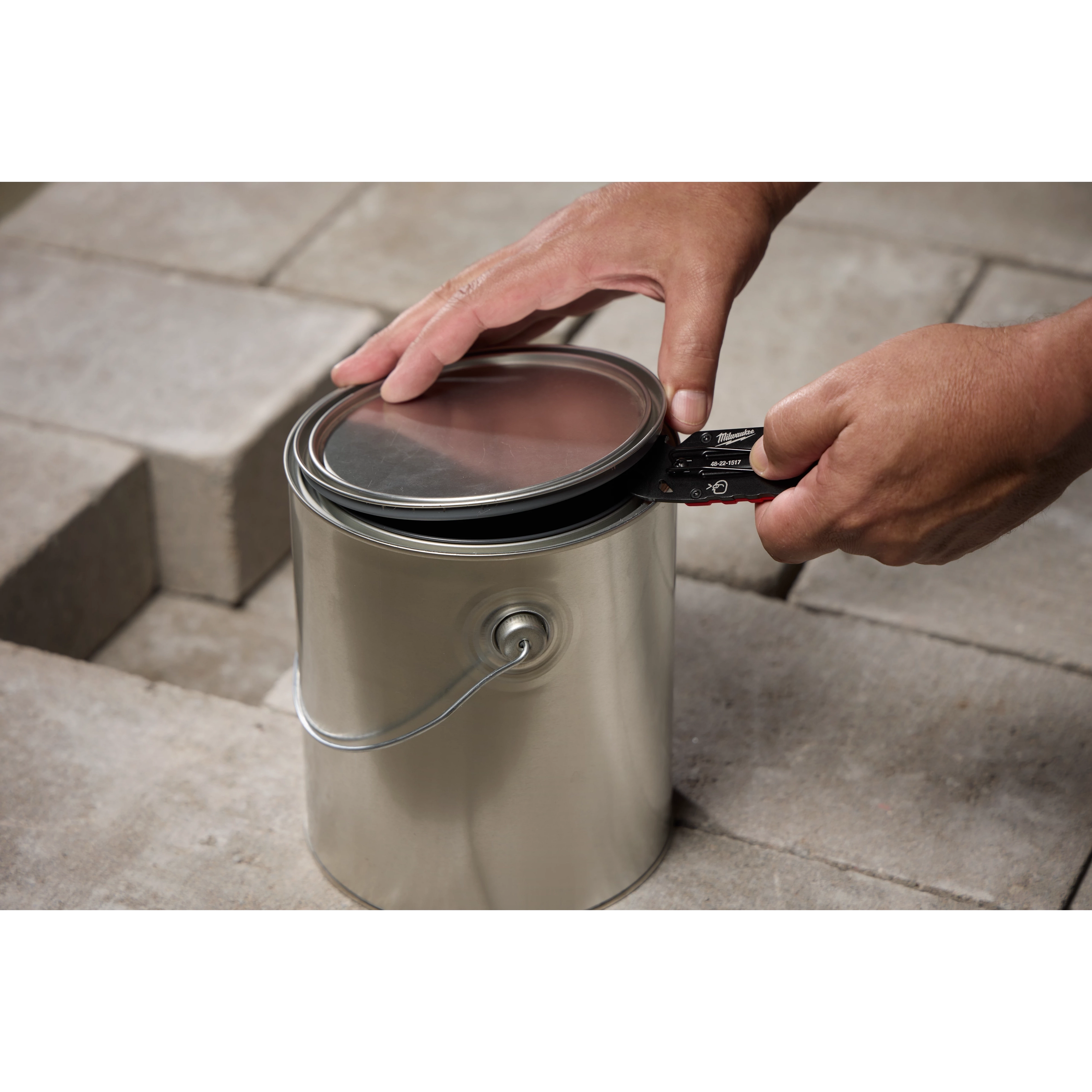 Hands using a Multi-function Compact Utility Knife to open the lid of a silver paint can. The knife is black with red accents and is being used to pry the lid open. Stone tiles are visible in the background.