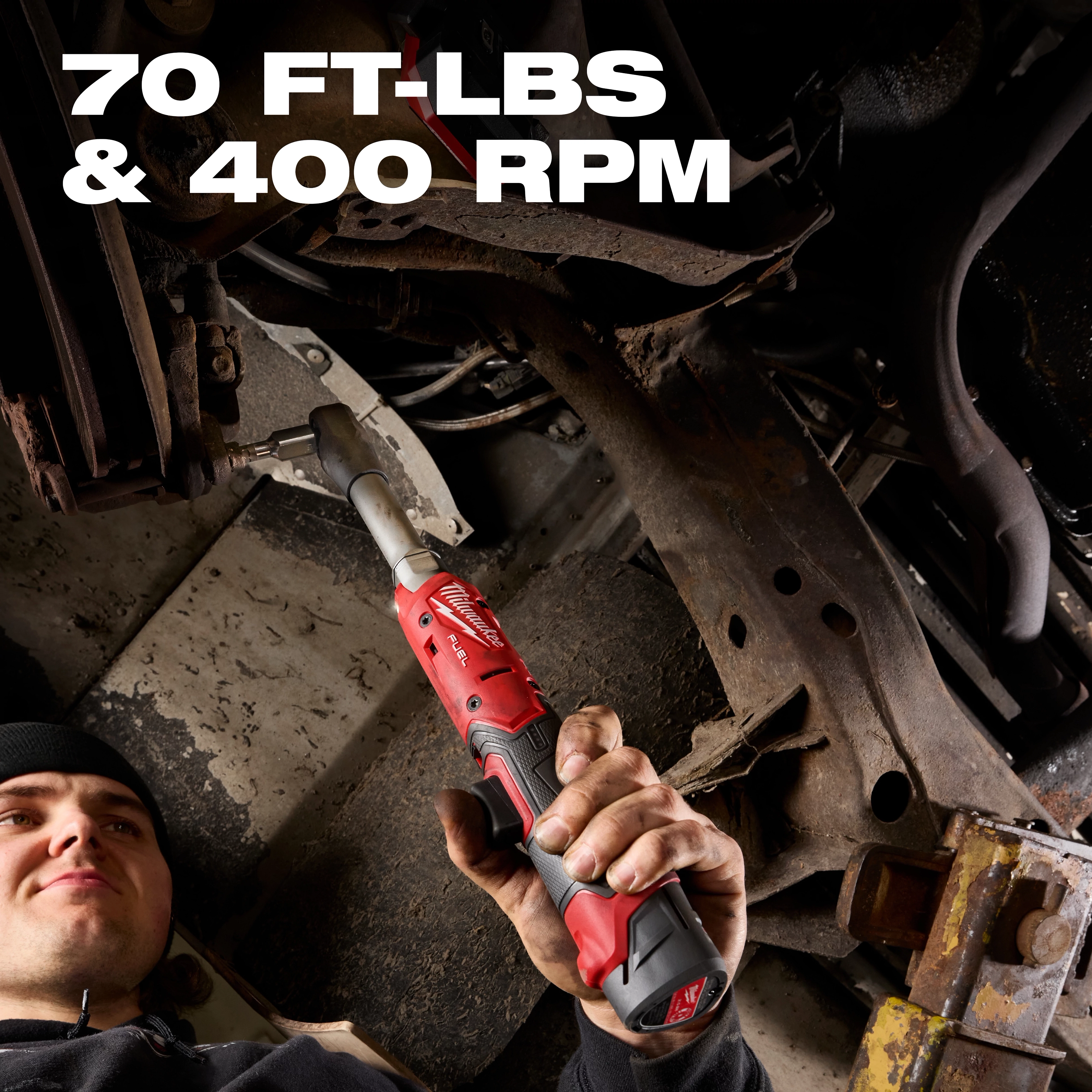 Hand holding an M12 FUEL 3/8" Extended Reach Ratchet working on metal parts under a vehicle frame.