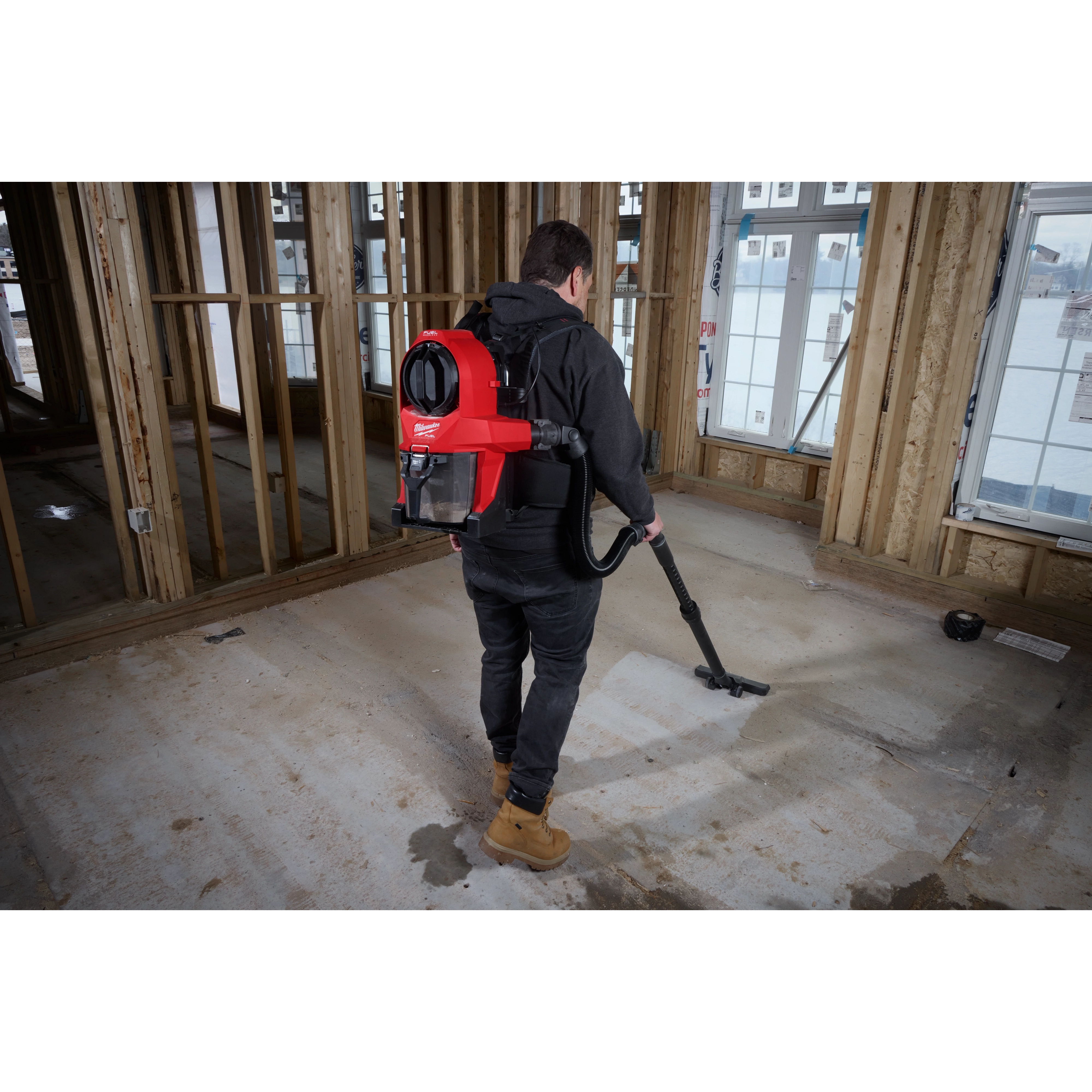 A person uses an M18 FUEL™ 3-in-1 Backpack Vacuum to clean a construction site. The individual wears the red backpack vacuum and operates the hose attachment to clean the floor in a partially framed building.