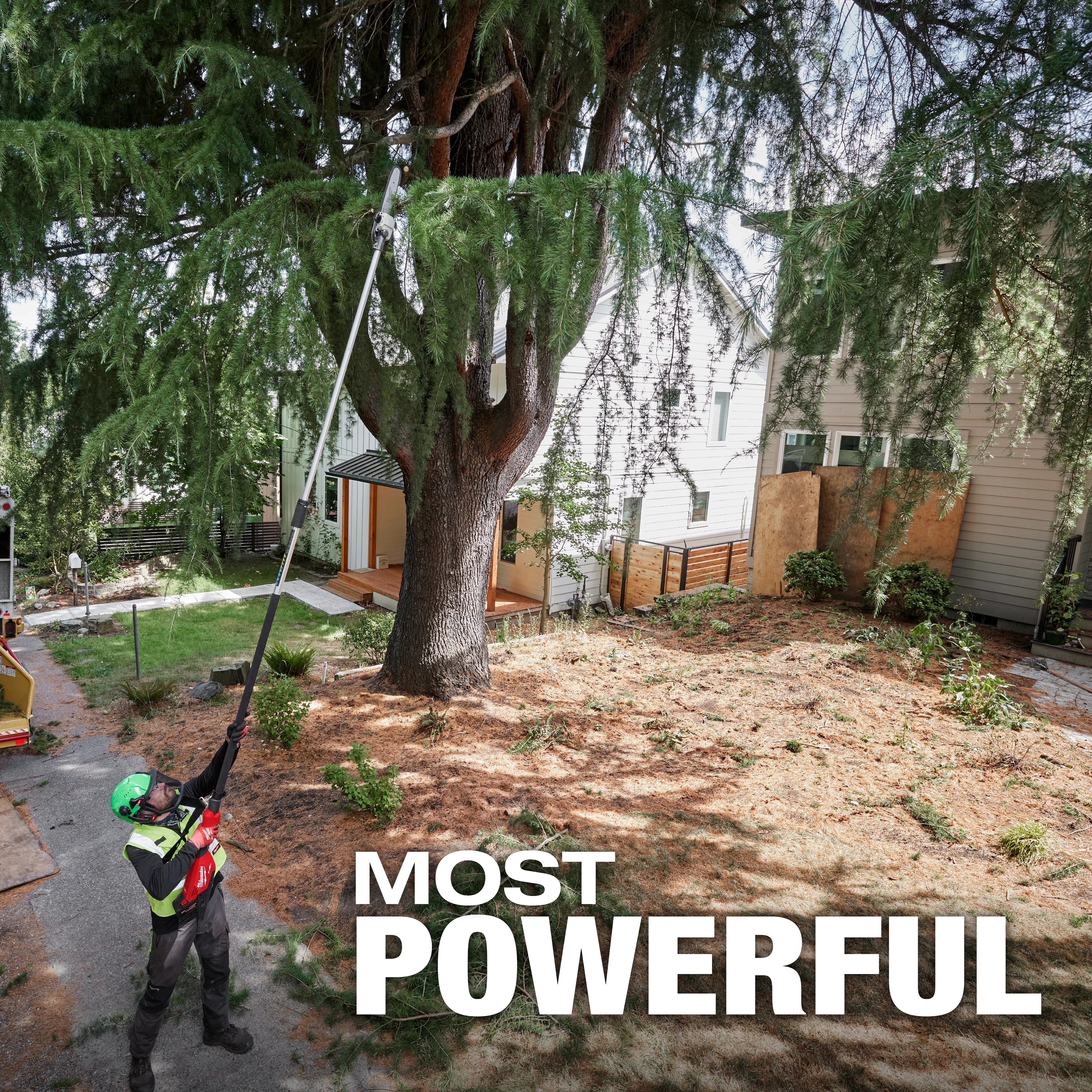 A worker uses the M18 FUEL™ Telescoping Pole Saw to trim branches high up in a large tree. The background shows a residential yard with houses, and the words "MOST POWERFUL" are prominently displayed at the bottom of the image.