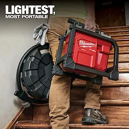 A person carries a Milwaukee Fuel portable air compressor up a wooden staircase. The product slogan "Lightest, Most Portable" appears in bold text at the top left of the image. The compressor is red with black casing and features ergonomic handles for easy transport.