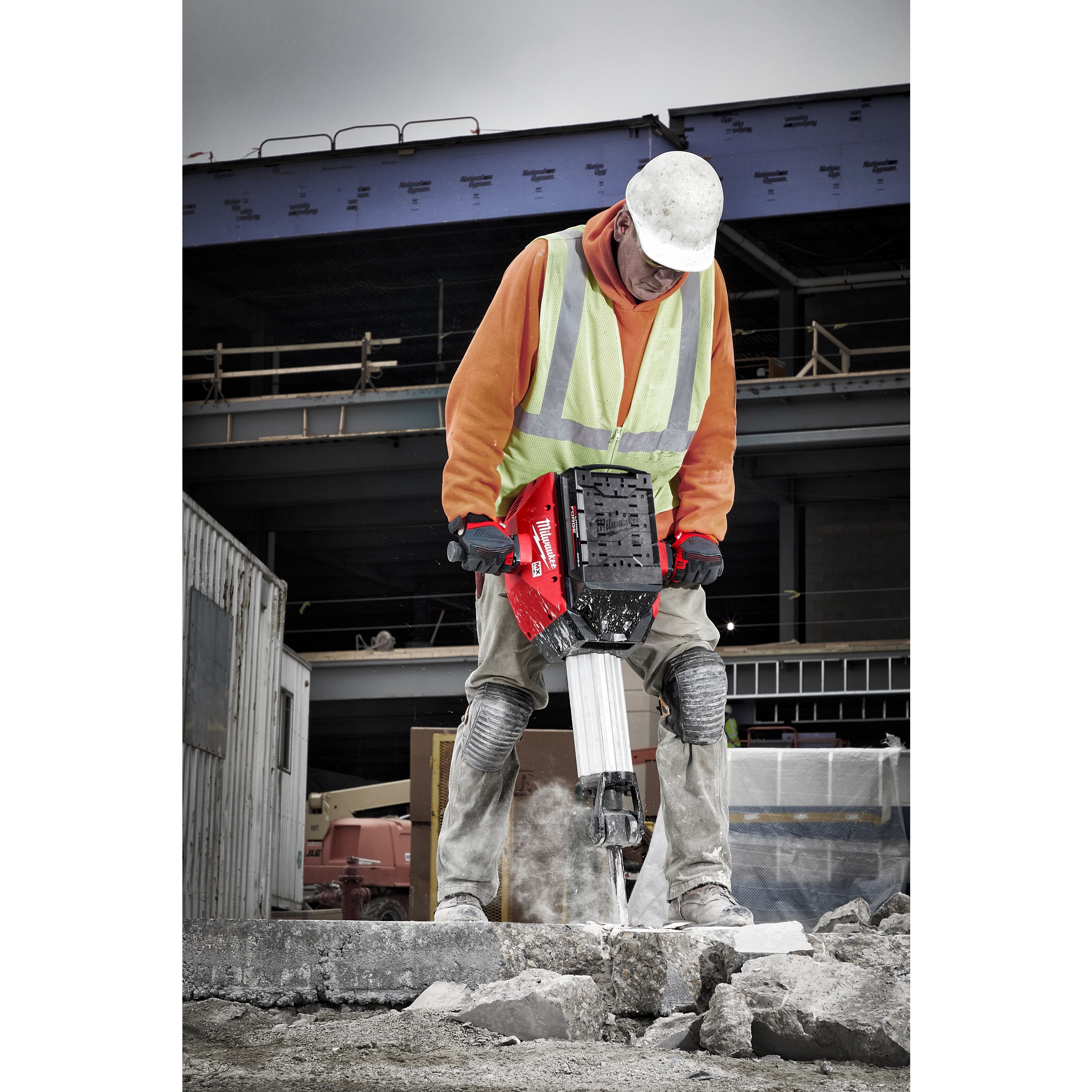 A person in construction gear operates an MX FUEL™ Breaker on a concrete surface at a building site. The breaker is red with black and white components, designed for heavy-duty demolition work. Structures and construction materials are visible in the background.