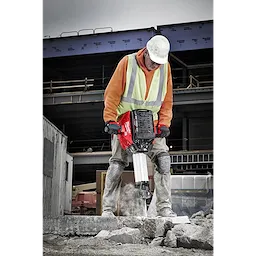 A person in construction gear operates an MX FUEL™ Breaker on a concrete surface at a building site. The breaker is red with black and white components, designed for heavy-duty demolition work. Structures and construction materials are visible in the background.