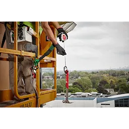 A worker uses the 5LB Quick-Connect Retractable Tool Lanyard to secure a heavy-duty wrench while standing on an elevated platform. The tool lanyard prevents tools from falling, enhancing safety. The background shows an urban landscape with buildings and greenery.