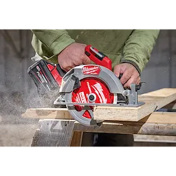 A person is using a red Milwaukee circular saw with a 7-1/4" 24T Thick Kerf Framing Circular Saw Blade to cut wood. Sawdust is visible from the cutting action. The circular saw has a black and red battery pack attached.