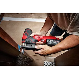 A person uses the Milwaukee M18 Fuel Jigsaw, Barrel Grip to cut wood. The red and black cordless jigsaw features an M18 lithium-ion battery and ergonomic design, providing precise control for detailed work. Dust and wood particles are visible as the tool operates.