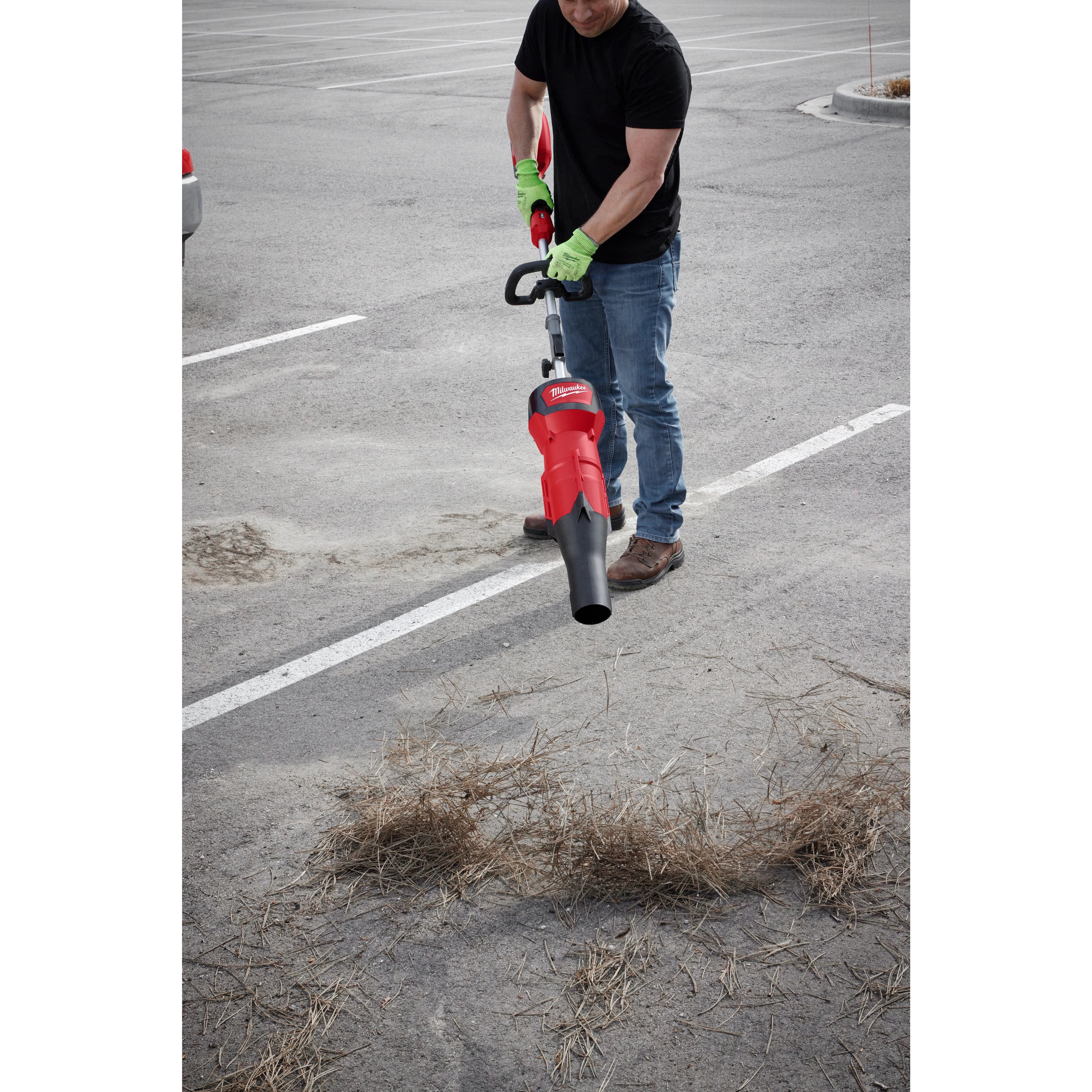 Man using Blower Attachment tool to clear straw and debris from a parking lot, wearing gloves, black shirt, and blue jeans.