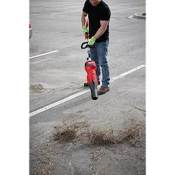 Man using Blower Attachment tool to clear straw and debris from a parking lot, wearing gloves, black shirt, and blue jeans.