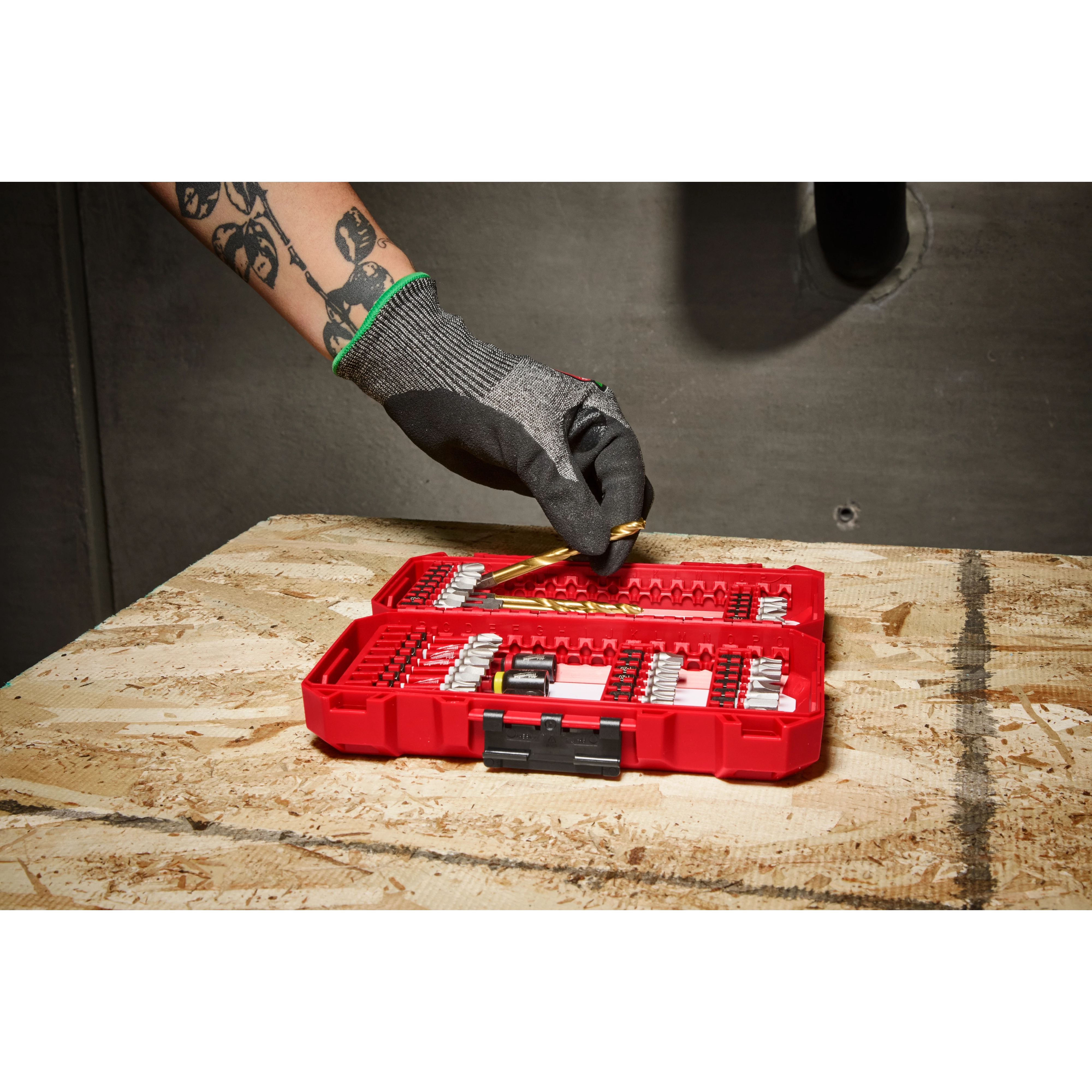 A person wearing gloves is picking a drill bit from an open red case of the SHOCKWAVE™ Impact Duty Drill & Drive Set – 50PC, which is placed on a wooden surface.