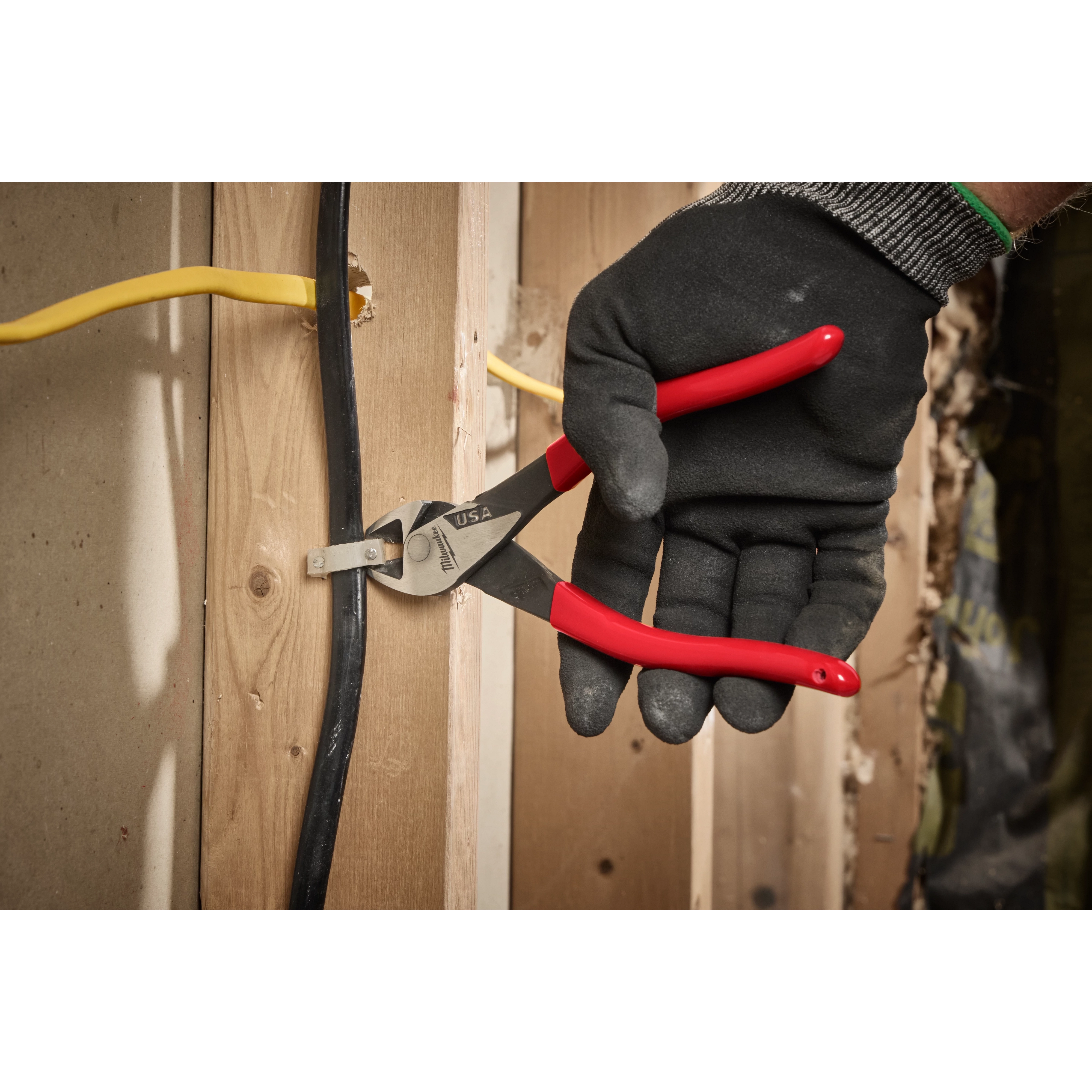 A person wearing a black glove uses ChannelLock wire cutters to trim a black cable fixed to a wooden stud. The wire cutters have red handles, and the cutting jaws grip the cable securely. There is also a yellow cable visible in the background.
