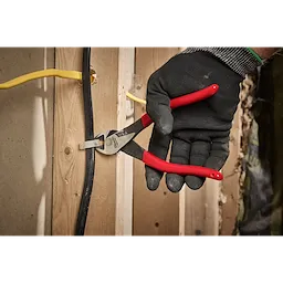 A person wearing a black glove uses ChannelLock wire cutters to trim a black cable fixed to a wooden stud. The wire cutters have red handles, and the cutting jaws grip the cable securely. There is also a yellow cable visible in the background.