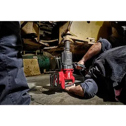 A person operates an M18 FUEL™ 1" D-Handle High Torque Impact Wrench w/ONE-KEY™ App Shot, working on the underside of a large machine. The red and black tool is in use on a bolt or attachment point, highlighting its industrial application.