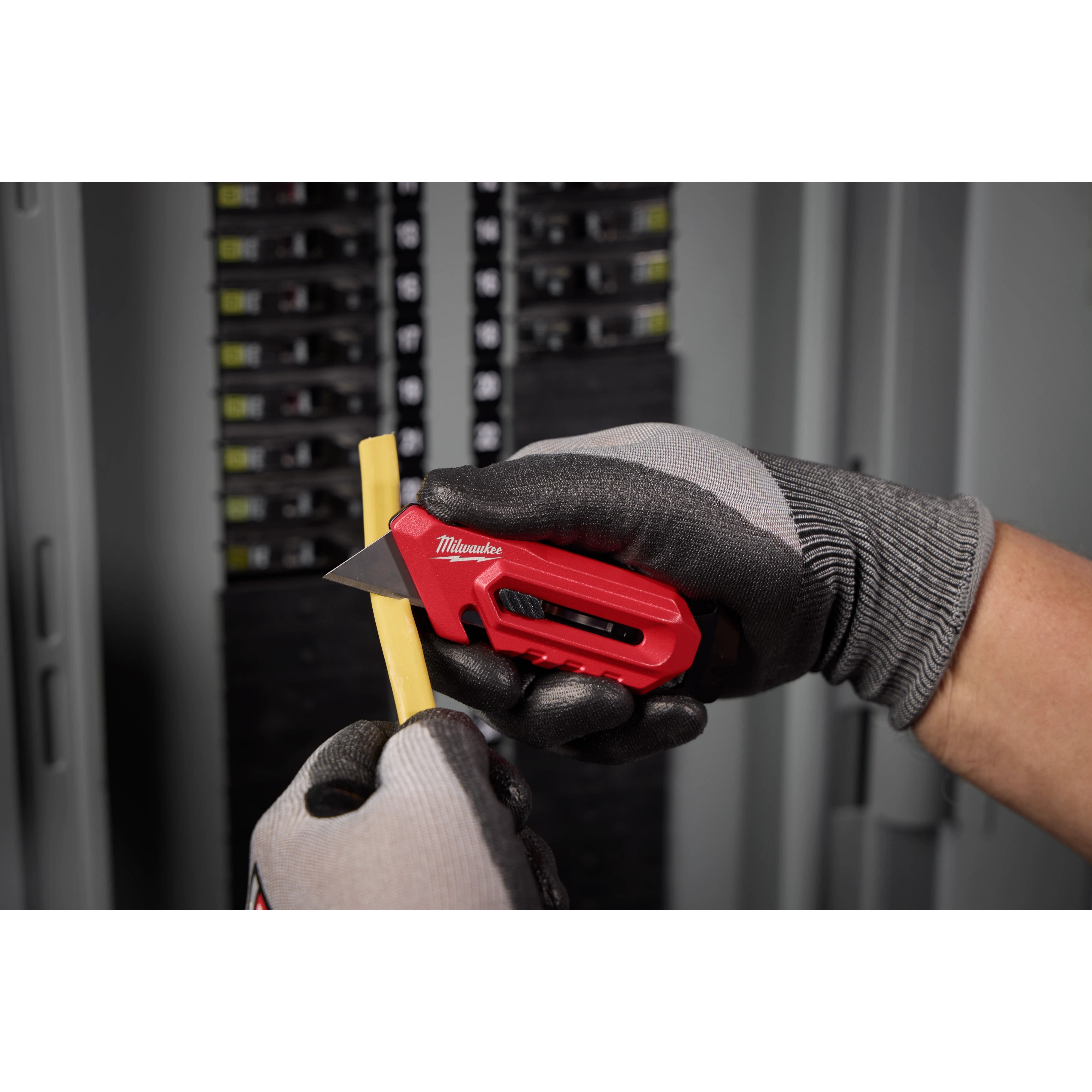 A person wearing gloves uses the Multi-function Compact Utility Knife to cut a yellow strip in front of an electrical panel. The knife has a red handle and a retractable blade, providing precise control for detailed tasks.