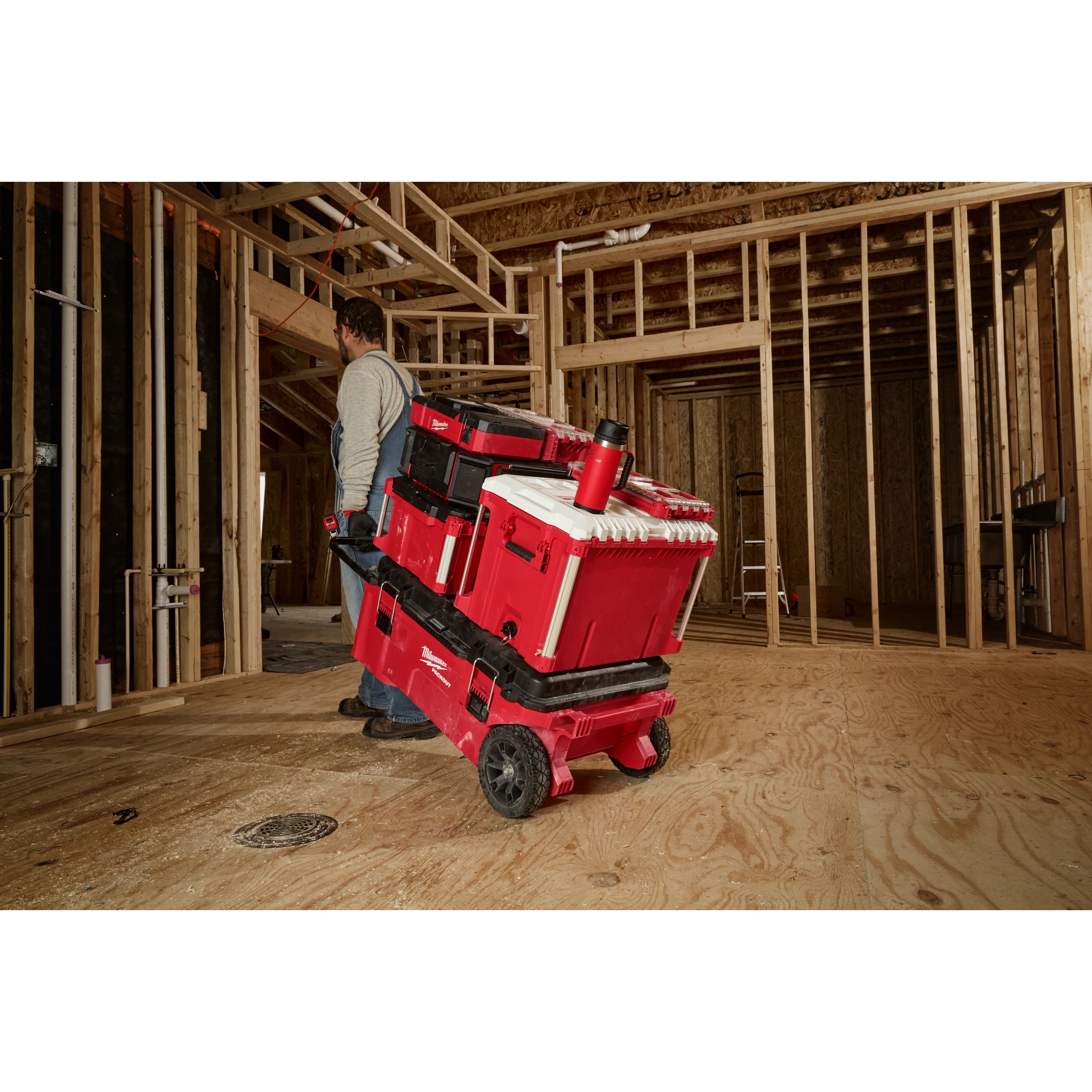 Man pulling a stack of red tool storage boxes on a cart in a construction site, with a PACKOUT 18oz Insulated Mug with Sip Lid on top.