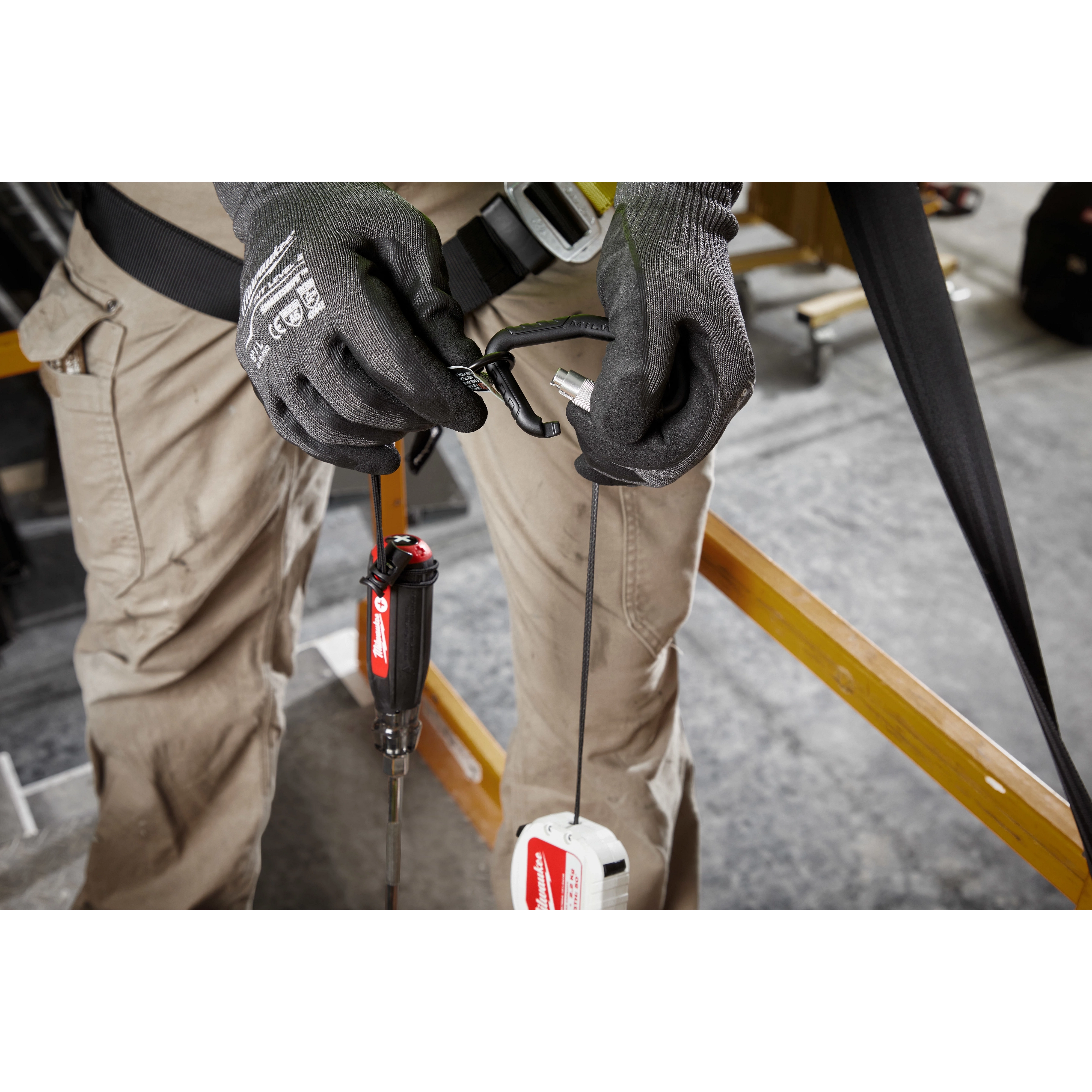 A worker attaches a tool to a 5LB Quick-Connect Retractable Tool Lanyard. The worker wears grey gloves and tan cargo pants, and they are standing in a workshop. The lanyard has a retractable feature for convenience and safety.