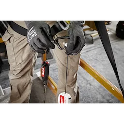 A worker attaches a tool to a 5LB Quick-Connect Retractable Tool Lanyard. The worker wears grey gloves and tan cargo pants, and they are standing in a workshop. The lanyard has a retractable feature for convenience and safety.