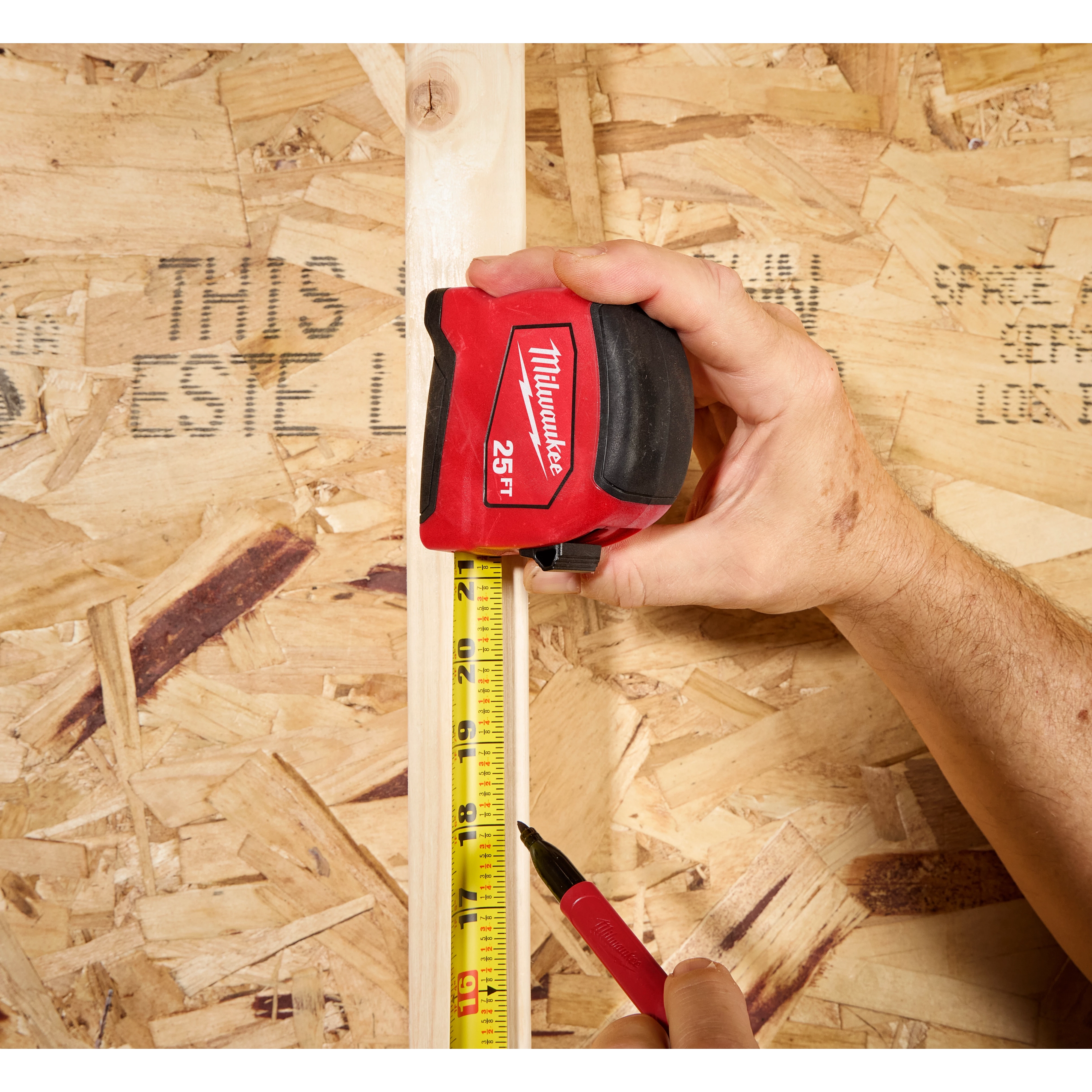 Hand holding a 25ft Tape Measure against a vertical board while marking the measurement with a pen.