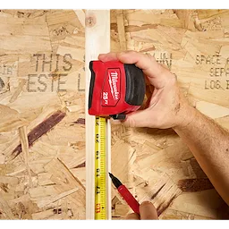 Hand holding a 25ft Tape Measure against a vertical board while marking the measurement with a pen.