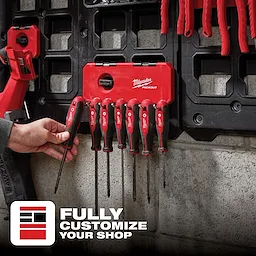 A person inserts a screwdriver into the red PACKOUT 7-Hook Rack, mounted on a workshop wall, with other tools hanging on it.