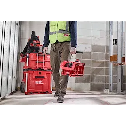 A person carrying a PACKOUT Compact Crate in a construction setting, with a stacked set of red PACKOUT toolboxes in the background.