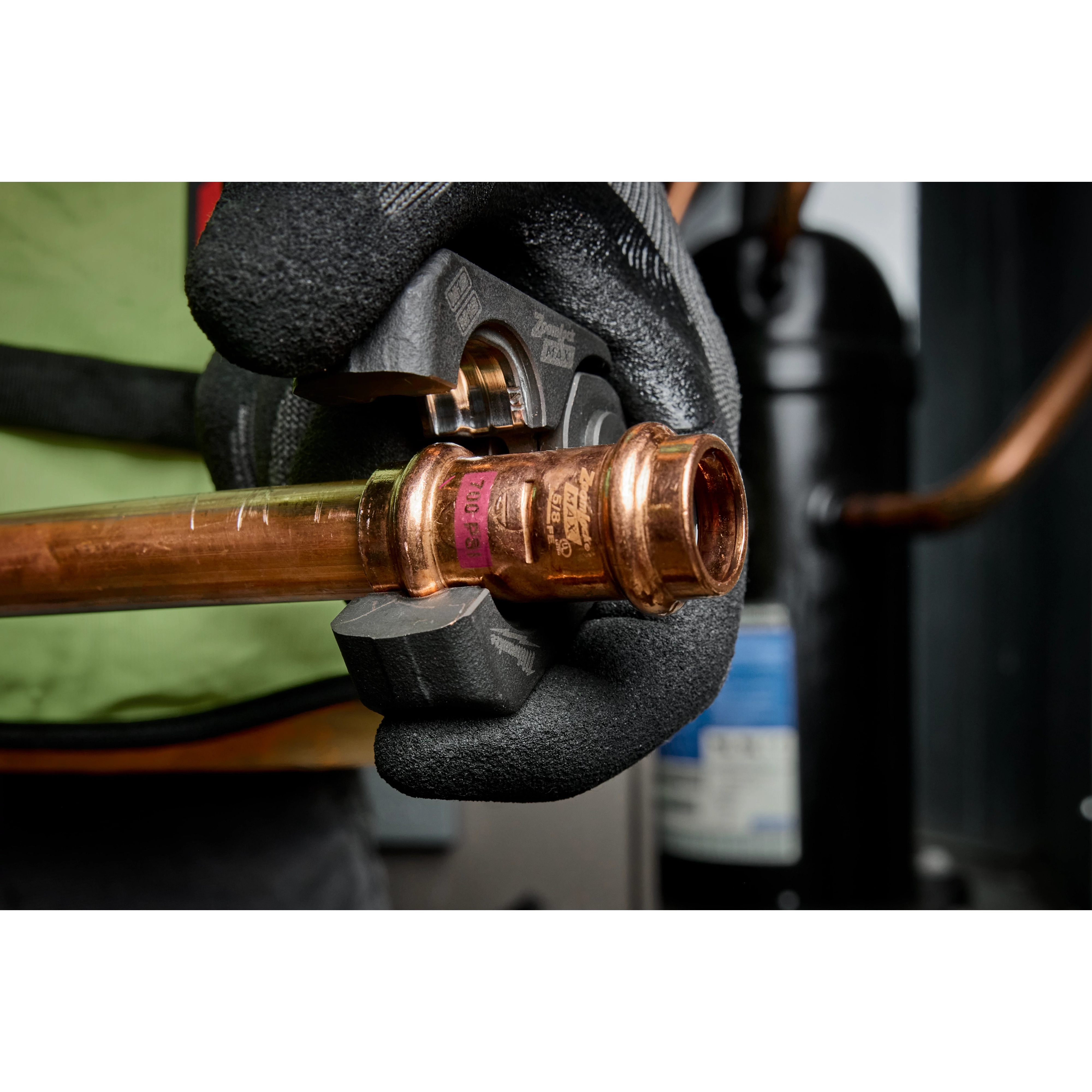 A person wearing black gloves uses the 1/4” – 7/8” ZoomLock MAX® & MaxiPro® Pivoting Press Ring Kit for M18™ FORCE LOGIC™ Press Tool to crimp a copper pipe fitting, demonstrating its application in plumbing tasks. The background shows industrial equipment.
