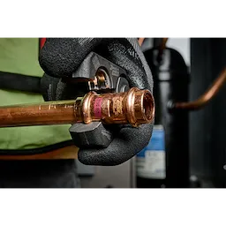 A person wearing black gloves uses the 1/4” – 7/8” ZoomLock MAX® & MaxiPro® Pivoting Press Ring Kit for M18™ FORCE LOGIC™ Press Tool to crimp a copper pipe fitting, demonstrating its application in plumbing tasks. The background shows industrial equipment.