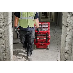 A construction worker in a green safety vest and black gloves is pulling a red cart filled with tools and materials through a narrow passage of a partially constructed building. The floor is covered with debris and electrical wires. The person’s face is not visible.