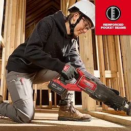 A woman wearing a black Women's FREEFLEX™ Pullover Hoodie and gray pants with reinforced pocket seams is working on a construction site, using a red power tool while kneeling. The scene is set inside a wooden framework of an unfinished building.