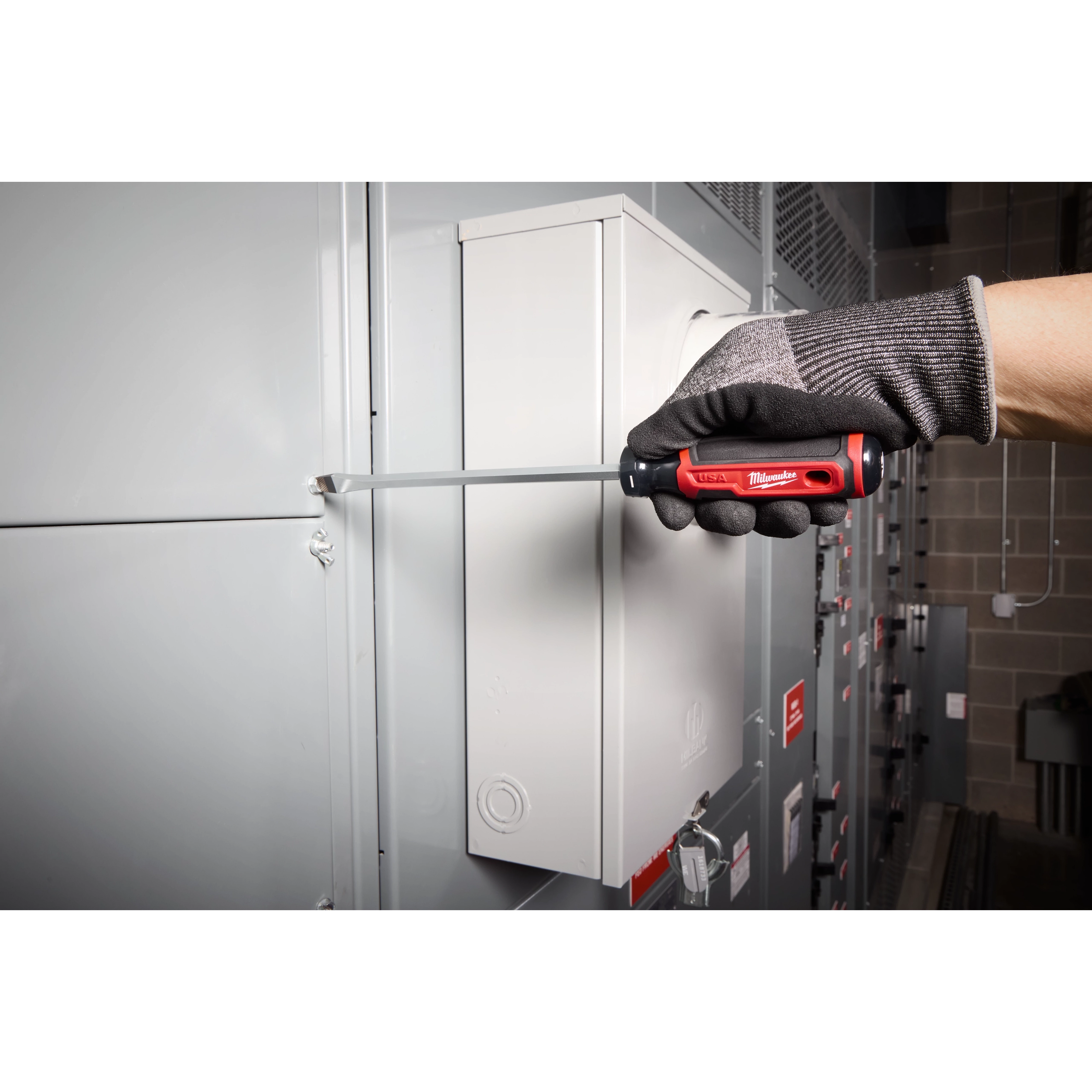 Gloved hand using 3/8" Slotted 8" Cushion Grip Screwdriver (USA) to turn a screw on a metal electrical panel.