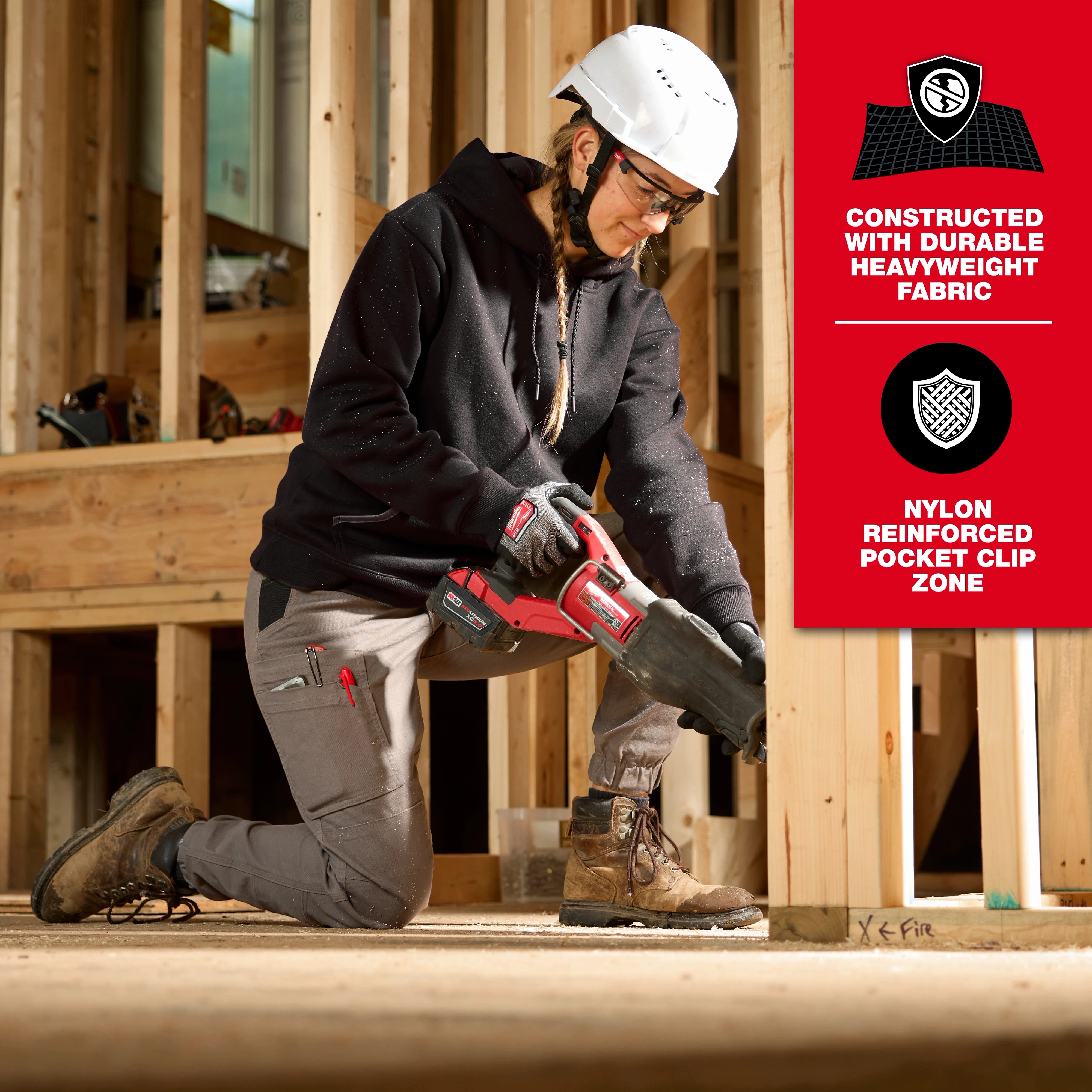 A woman wearing Women's GRIDIRON™ Double Knee Jogger Pants is kneeling on a wooden floor in a construction setting, holding a red power tool. The pants are highlighted for their durable heavyweight fabric and nylon reinforced pocket clip zone. Safety gear includes a white helmet and work boots.