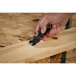 A person uses a Milwaukee Fastback Flip Utility Knife to cut green strapping wrapped around a wooden board. The black and red knife is designed for easy handling and efficient cutting. The hand technique demonstrates the versatility of the utility knife on construction materials.