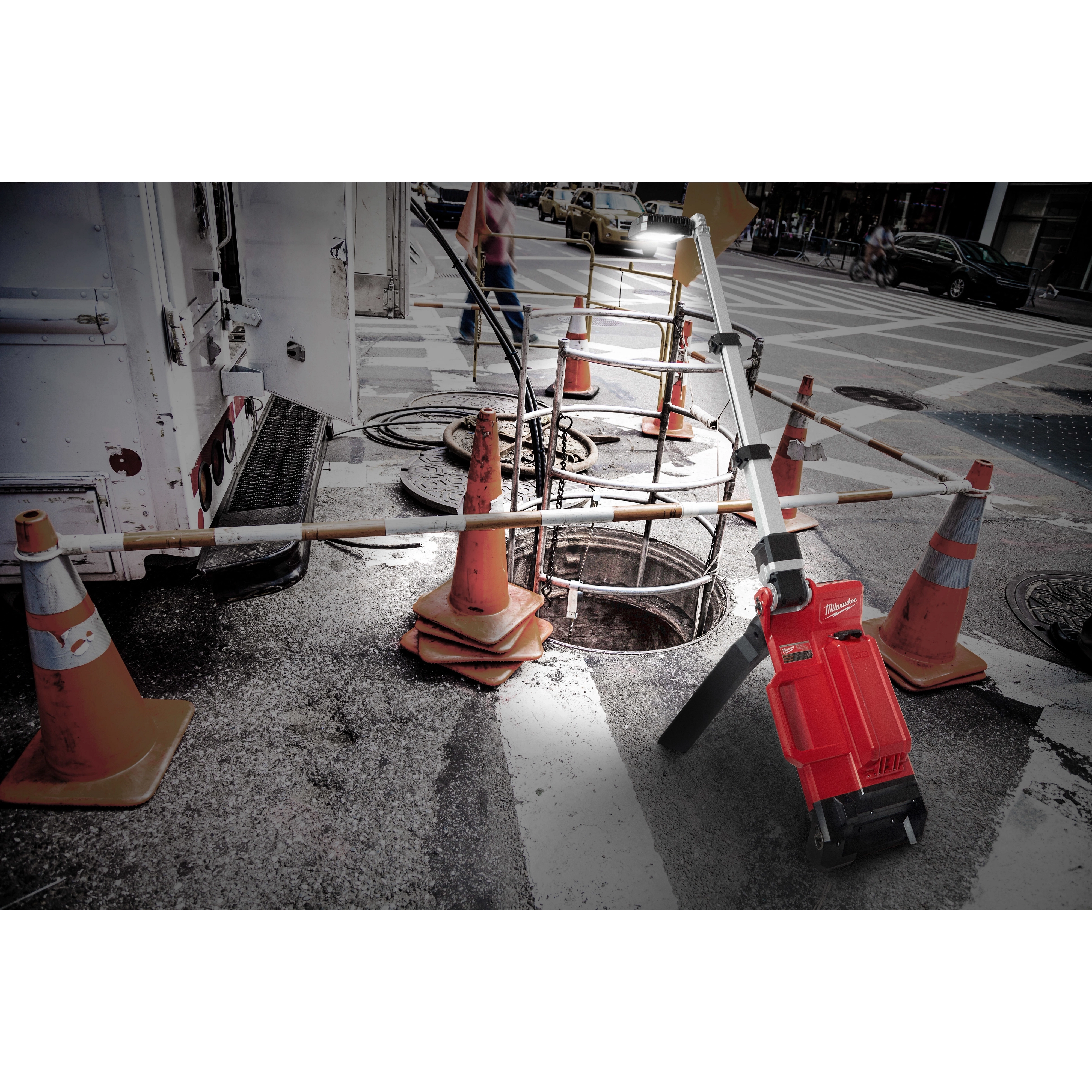 M18%20DUAL%20PACK%20TOWER%20LIGHT%20W%2FONE%20KEY is positioned near construction cones, illuminating an open manhole on a city street at dusk.