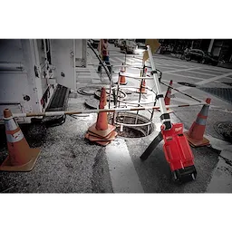 M18%20DUAL%20PACK%20TOWER%20LIGHT%20W%2FONE%20KEY is positioned near construction cones, illuminating an open manhole on a city street at dusk.