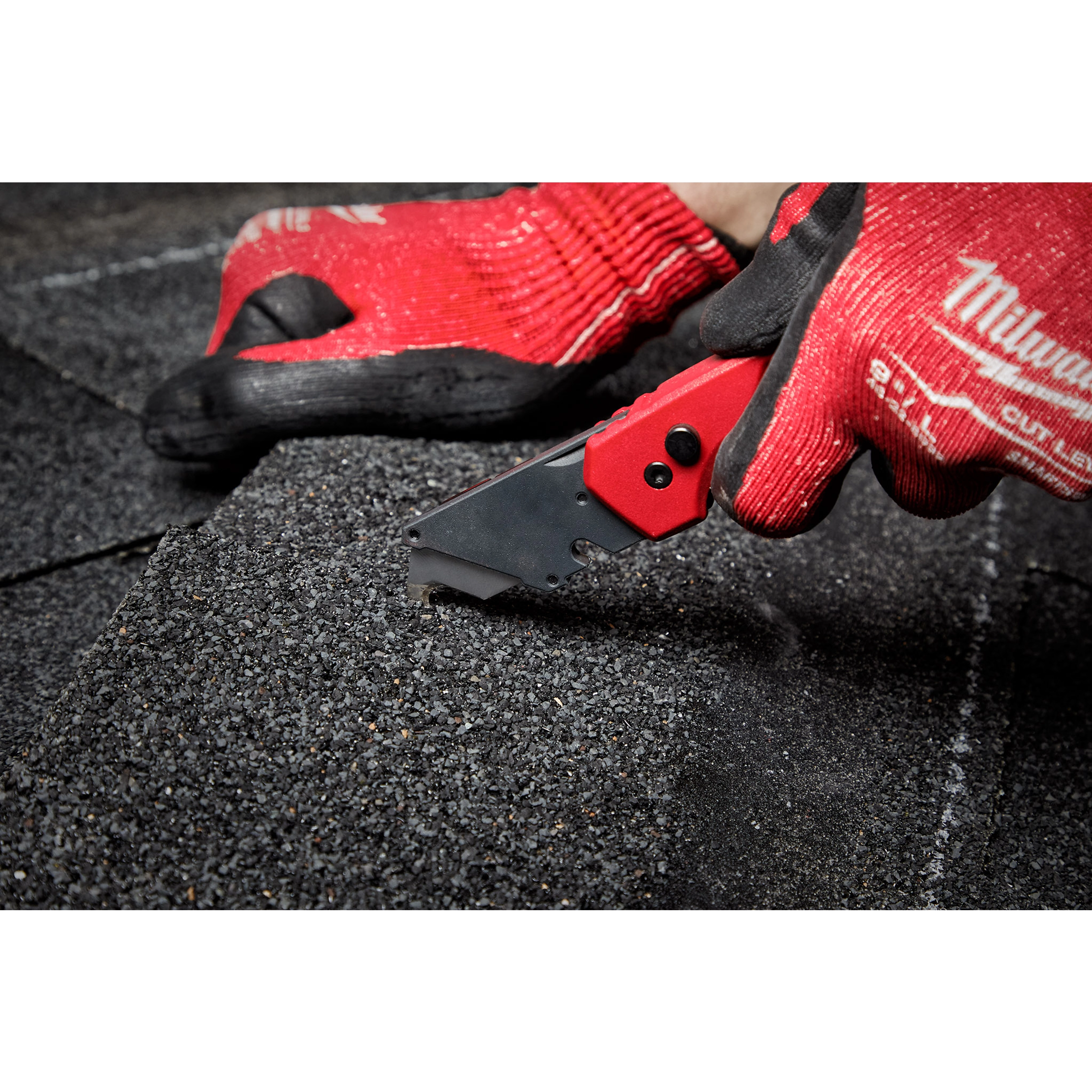 A person wearing red gloves is cutting through shingles with the FASTBACK Folding Utility Knife on a dark, rough surface.