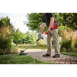 A man uses the M18™ Brushless String Trimmer to edge a lawn beside a brick walkway. The trimmer features a long handle for better reach and a red-colored motor casing, highlighting its battery-operated design. Dense greenery and tall grass surround the path in an outdoor setting.