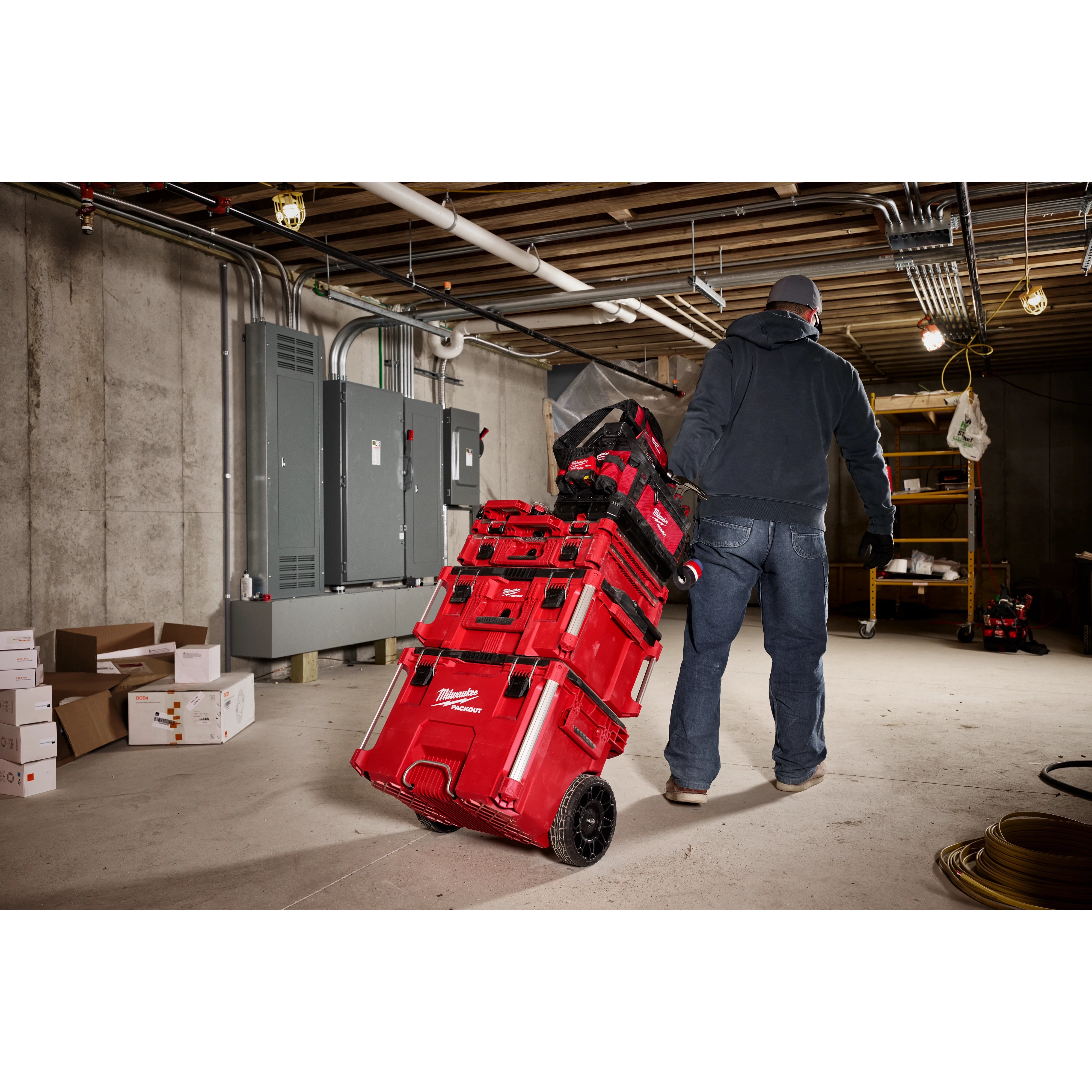 A person is pulling a red PACKOUT™ 15" Structured Tote system with multiple stacked toolboxes on wheels in an industrial storage room. The room has concrete walls, electrical panels, and visible piping and wiring on the ceiling. Other boxes and equipment are seen in the background.
