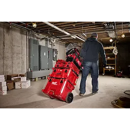 A person is pulling a red PACKOUT™ 15" Structured Tote system with multiple stacked toolboxes on wheels in an industrial storage room. The room has concrete walls, electrical panels, and visible piping and wiring on the ceiling. Other boxes and equipment are seen in the background.