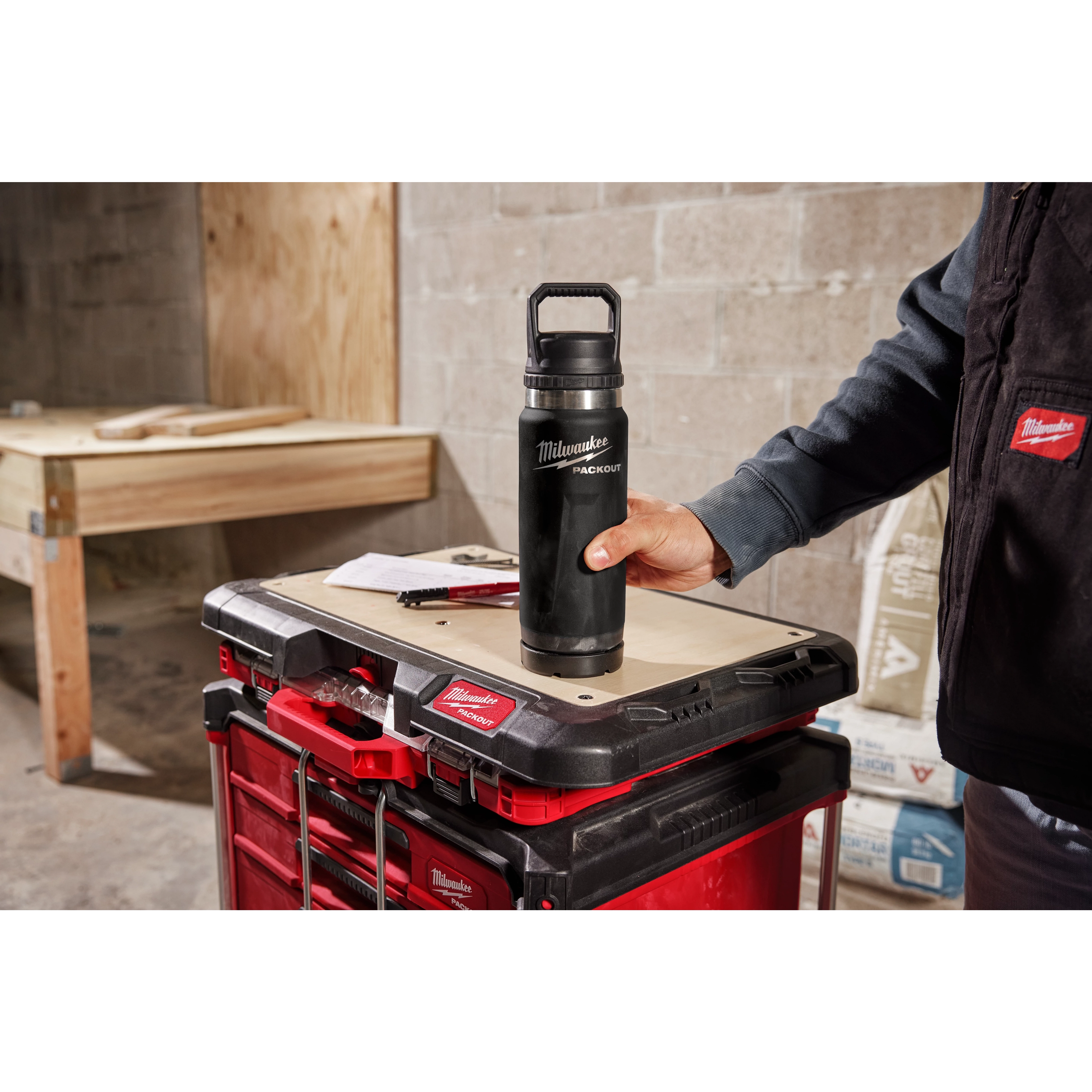 A person holds the PACKOUT 24oz Insulated Bottle with Chug Lid on a workbench adorned with red and black toolboxes in a workshop.