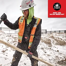 Man wearing FREEFLEX Insulated Bib Overalls, wind and water resistant, with a safety vest and helmet, working in snowy conditions.